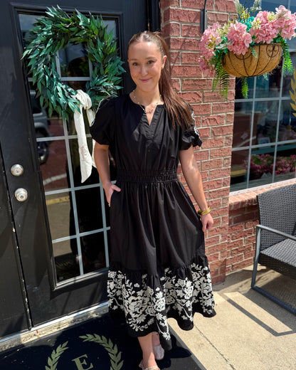 The Tracey Aven Vines Black Dress is an embroidered black dress with white floral detailing at the hem and a smocked waist. Available in sizes XS–XL.