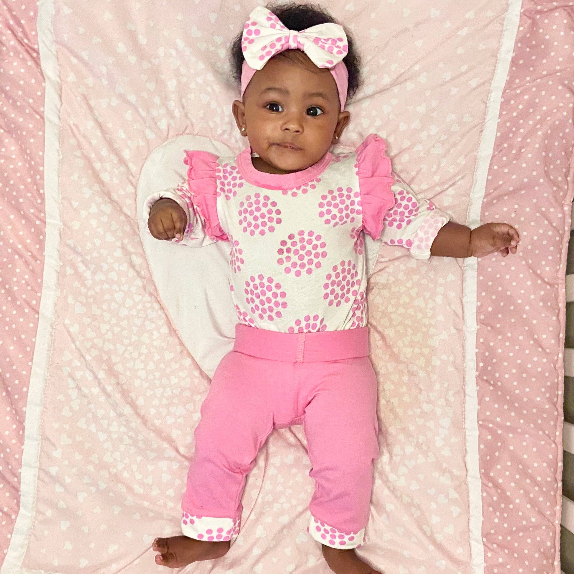 A baby girl wearing a Layette Girls Baby clothes Shower Gift Pink Polka Dot Easter outfit with polka dots and a matching bow headband lies on a pink patterned blanket made of Egyptian cotton, looking up with a curious expression.