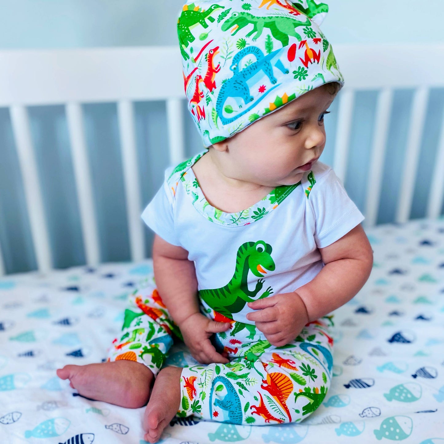 Baby moedling the set in a white crib in a blue room. Sitting and looking to the right.