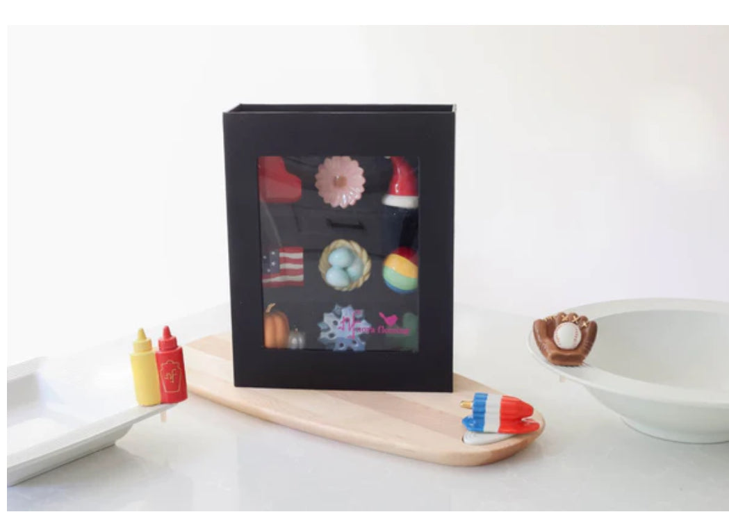 **Product: Keepsake Box**

- **Features:**
  - Holds 9 items
  - Transparent front
  
- **Contents:**
  - Colorful assorted-themed pins/magnets (holiday items, patriotic symbols)
  - Minis of condiment bottles, small baseball glove, popsicle-shaped items

- **Design:**
  - White and wooden surface