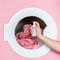 Sweet Grace Stain Remover spray bottle held over laundry inside a washing machine door. Available in 8oz and 16oz sizes.