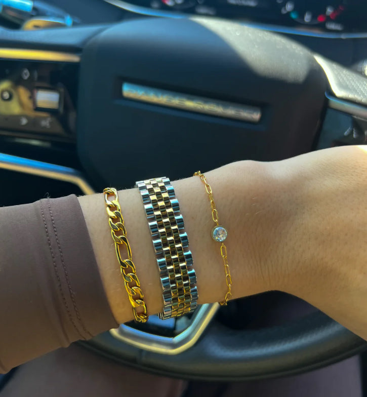 A wrist adorned with three bracelets: chunky gold chain; two-tone metal The Shelby Bracelet; delicate gold chain with small round gemstone. Available sizes: Small, Medium, Large.