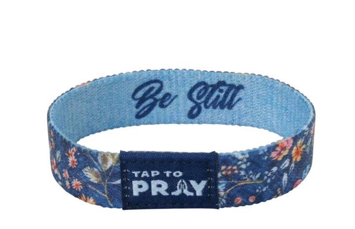 Tap To Pray™ Wristband in Wildflower. Features an elastic floral design, an inner “Be Still” inscription, and a dark blue front tab with a “Tap To Pray” label and praying hands icon.