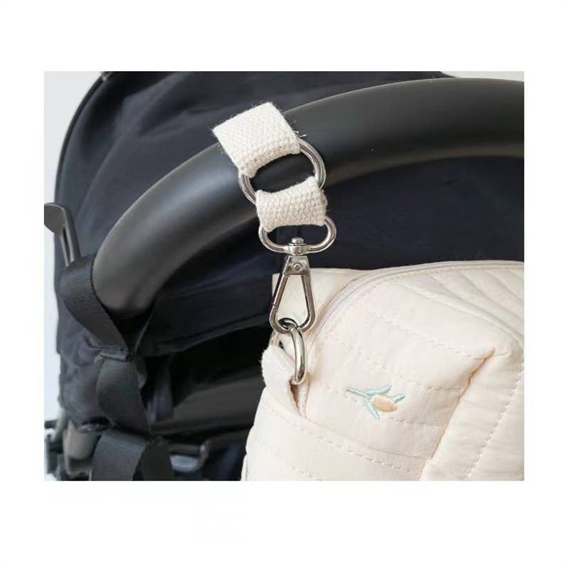 Close-up of a stroller handle with a velcro strap attached to a metal carabiner that is clipped to an Olive Embroidered Stroller and Diaper Bag with a small embroidered bluebird.