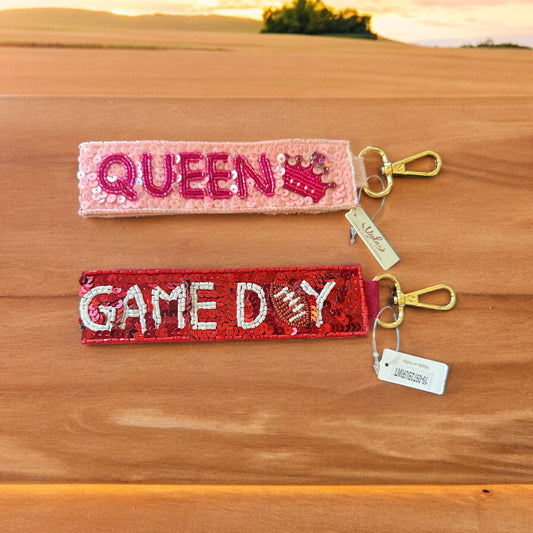 Two vibrant Sequin Wrist Key Holders. Pink one says "QUEEN" with a crown design, red one says "GAME DAY" with a football design. Each has a gold clasp and small tag. Size: Standard wristlet size.