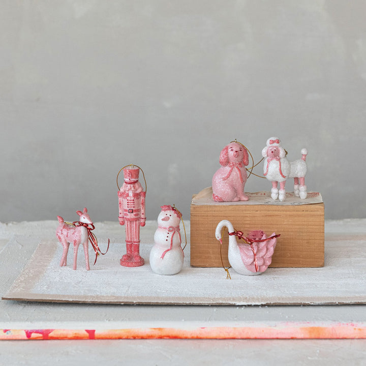 Festive pink and white resin Christmas ornaments: deer, nutcracker, snowman, two poodles, and a swan, each with a gold hanging string.