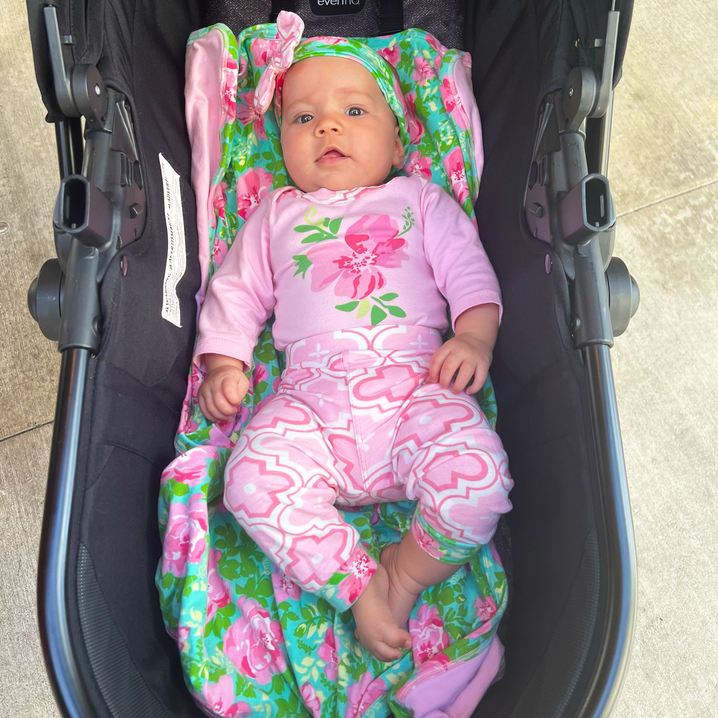 A baby in a Pink Floral Blanket+Bib Baby Shower Gift Set outfit is lying in a black stroller covered with an Egyptian cotton blanket, looking upwards with a curious expression. The baby appears relaxed and comfortable.