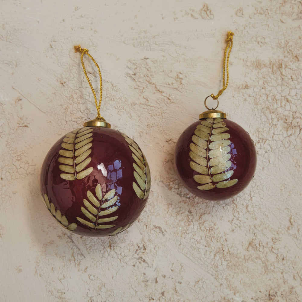 Two round, dark red glass ball ornaments with embedded eucalyptus leaves and gold leaf fern patterns, including gold hanging strings.