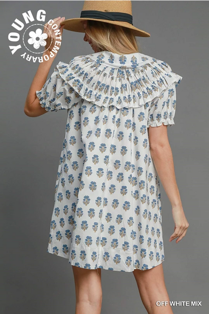 Loose-fitting white cotton mini dress with blue and yellow florals, pleated collar, puff sleeves. Sizes: XS–XL.
