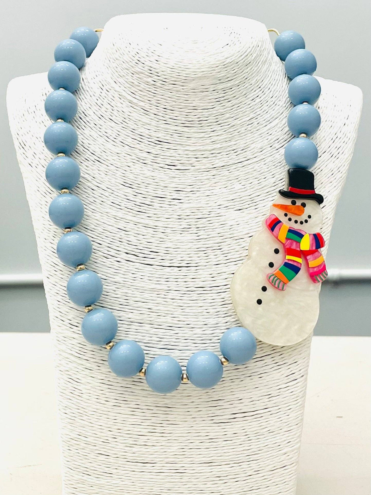 HOLIDAY - Beaded Snowman Necklace (blue/white) BNK1216 features large light blue beads and a snowman pendant with a black hat and rainbow-striped scarf. Available in multiple sizes.