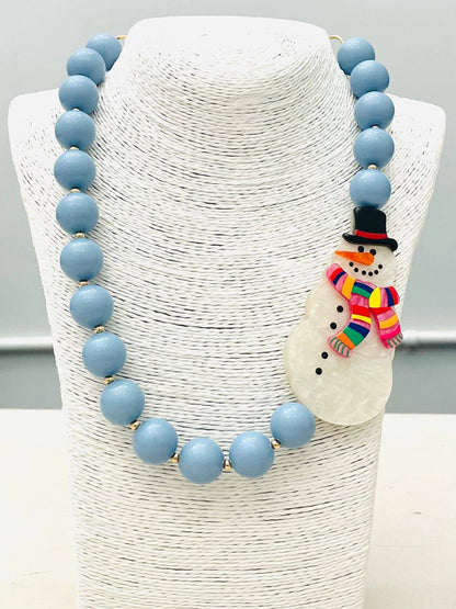HOLIDAY - Beaded Snowman Necklace (blue/white) BNK1216 features large light blue beads and a snowman pendant with a black hat and rainbow-striped scarf. Available in multiple sizes.
