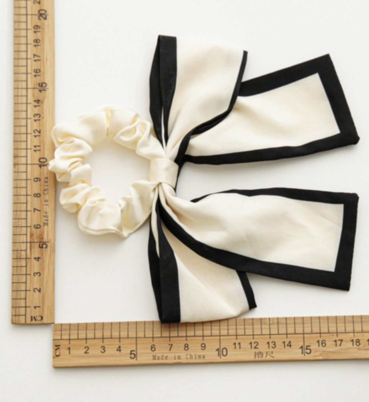 Two bow hair ties in cream and black colors. Crafted with robust and high-quality material, providing durability and style. The dimensions as shown next to a ruler, allows easy understanding of size compatibility. Suitable for all hair types. Available in one standard size.