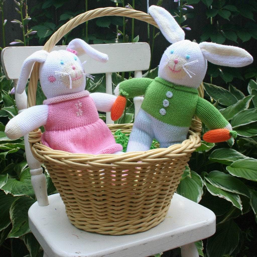 Two Harriett the Bunny Knit Doll: 12" Plush in a wicker basket, one wearing a pink dress and the other in a green sweater, sitting outdoors on a white chair with greenery in the background.