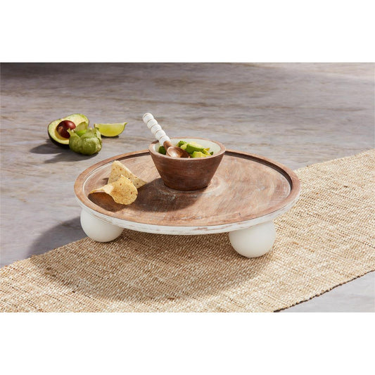 The Pedestal Chip N Dip is a round mango wood tray with white feet, featuring a small wooden bowl for dips. Sizes available: One size.