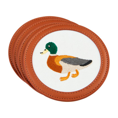 The Embroidered Icon Coaster Sets feature four round leather coasters adorned with needlepoint embroidery images of a mallard duck on a white fabric background. The duck is detailed with a green head, brown body, and yellow legs. Trimmed with faux leather for an elegant finish. Available in 3 styles.