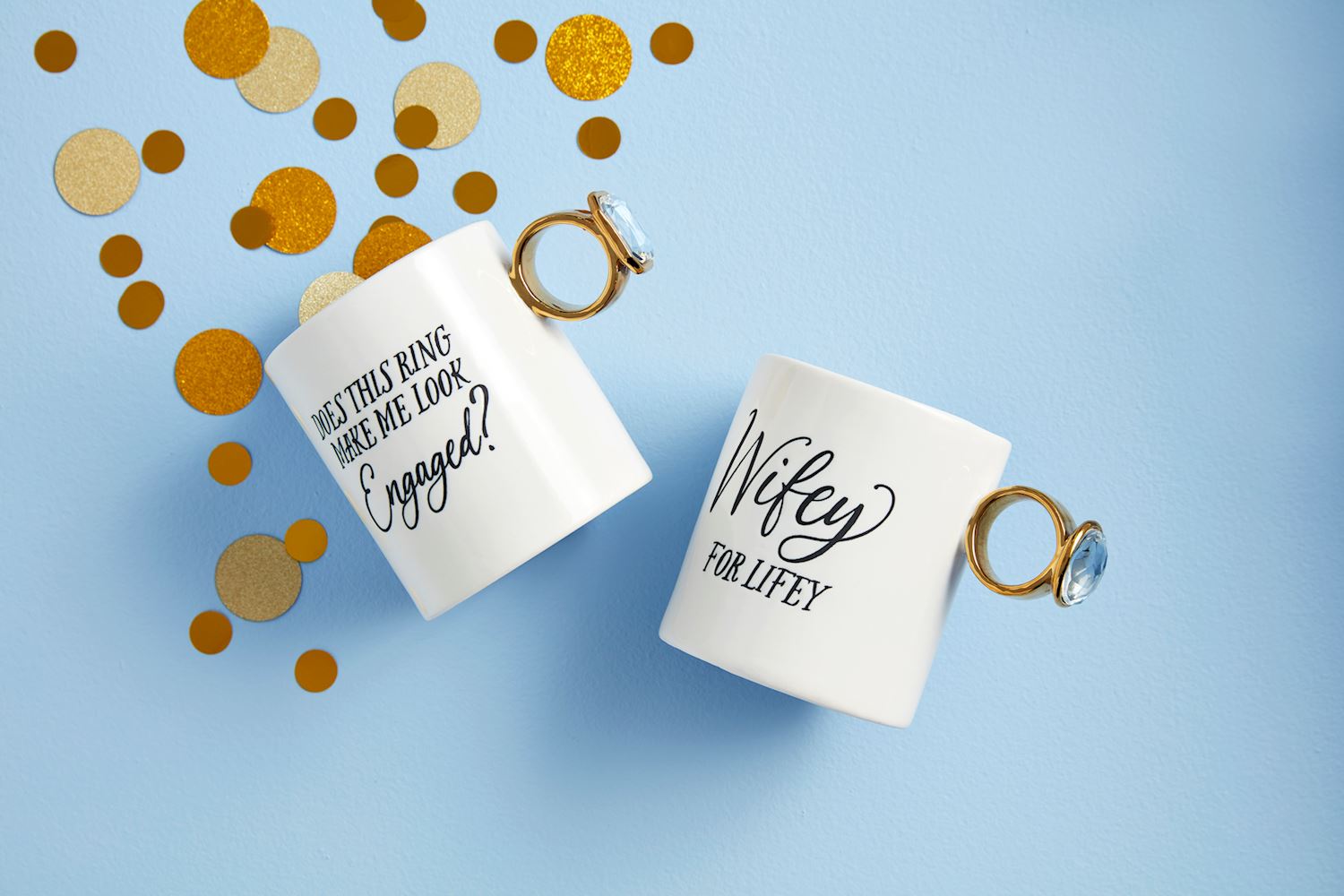 The Bridal Ring Mug set features two white mugs with ring-shaped handles. One mug reads "Does this ring make me look engaged?" and the other says "Wifey for Lifey," decorated with gold and brown confetti on a light blue background. Sizes: Standard mug size.