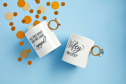 The Bridal Ring Mug set features two white mugs with ring-shaped handles. One mug reads "Does this ring make me look engaged?" and the other says "Wifey for Lifey," decorated with gold and brown confetti on a light blue background. Sizes: Standard mug size.