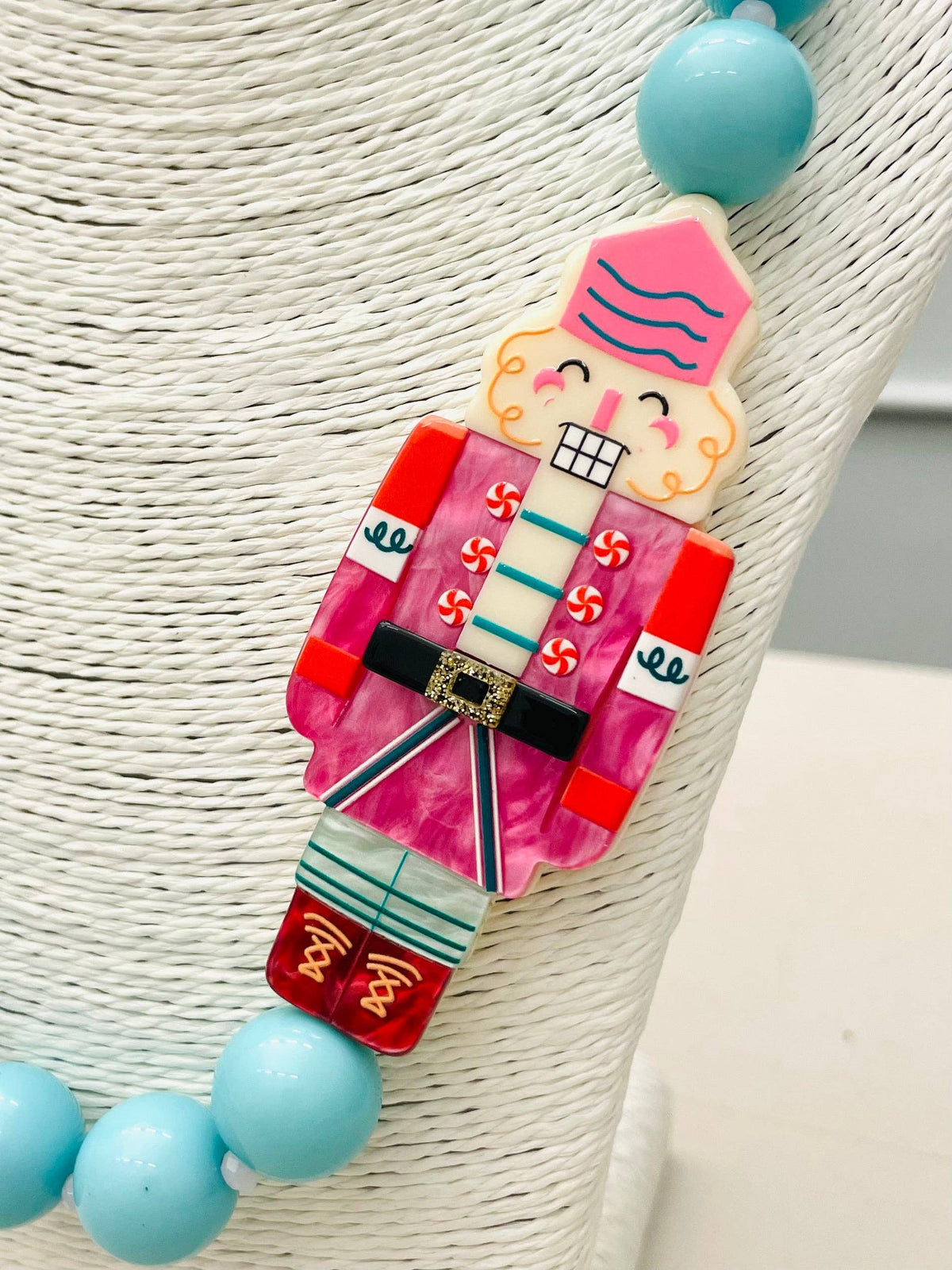 The HOLIDAY - Beaded Nutcracker Necklace (blue/pink) BNK1214 features a Nutcracker pendant with a pink hat, red jacket, and blue stripes on a necklace with large blue beads. Available in multiple sizes.