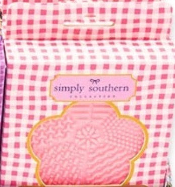 Pink and white checkered box labeled “Makeup Brush Cleanser” with a flower-shaped cutout showing a textured pink silicone cleaning pad inside; sizes available: not specified.