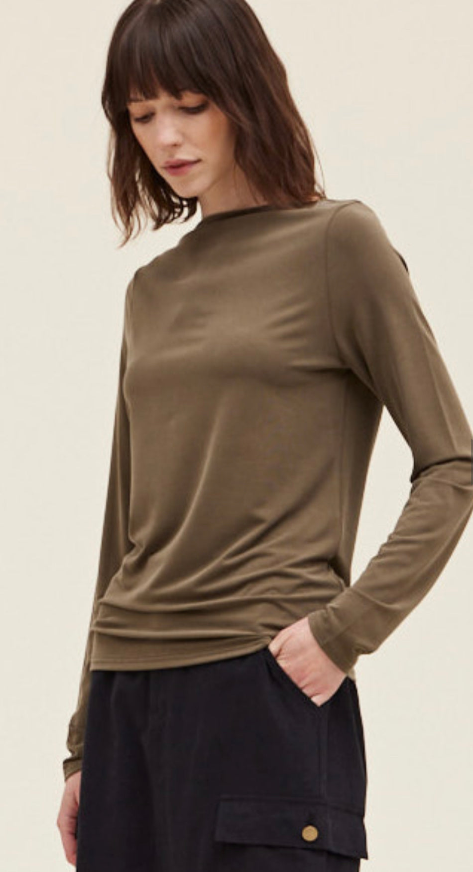 A woman with shoulder-length dark hair wearing a fitted Dusty Olive Tee made of polyester and standing against a pale background, looking downward with her hand in her pocket.