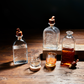 Four embossed glass decanters, one half-filled with amber liquid and three empty with cork stoppers. Two empty glasses, one marked "LOVE IS MESSY // BUY WHISKEY," sit in front. Sizes: Standard decanter size.