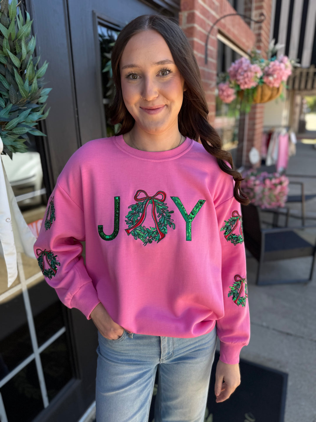 Pink Sarah Joy Wreath Sweatshirt with “JOY” graphic, sequined wreath and red bow detail, and wreath sleeve accents. Available in all sizes.