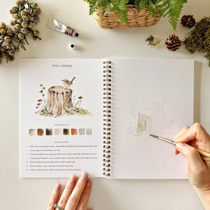 A hand sketches a tree stump with a bird in the Woodland Watercolor Workbook. The left page shows a painted stump, color palette, painting instructions, and pinecones. Available sizes for the workbook are not shown.