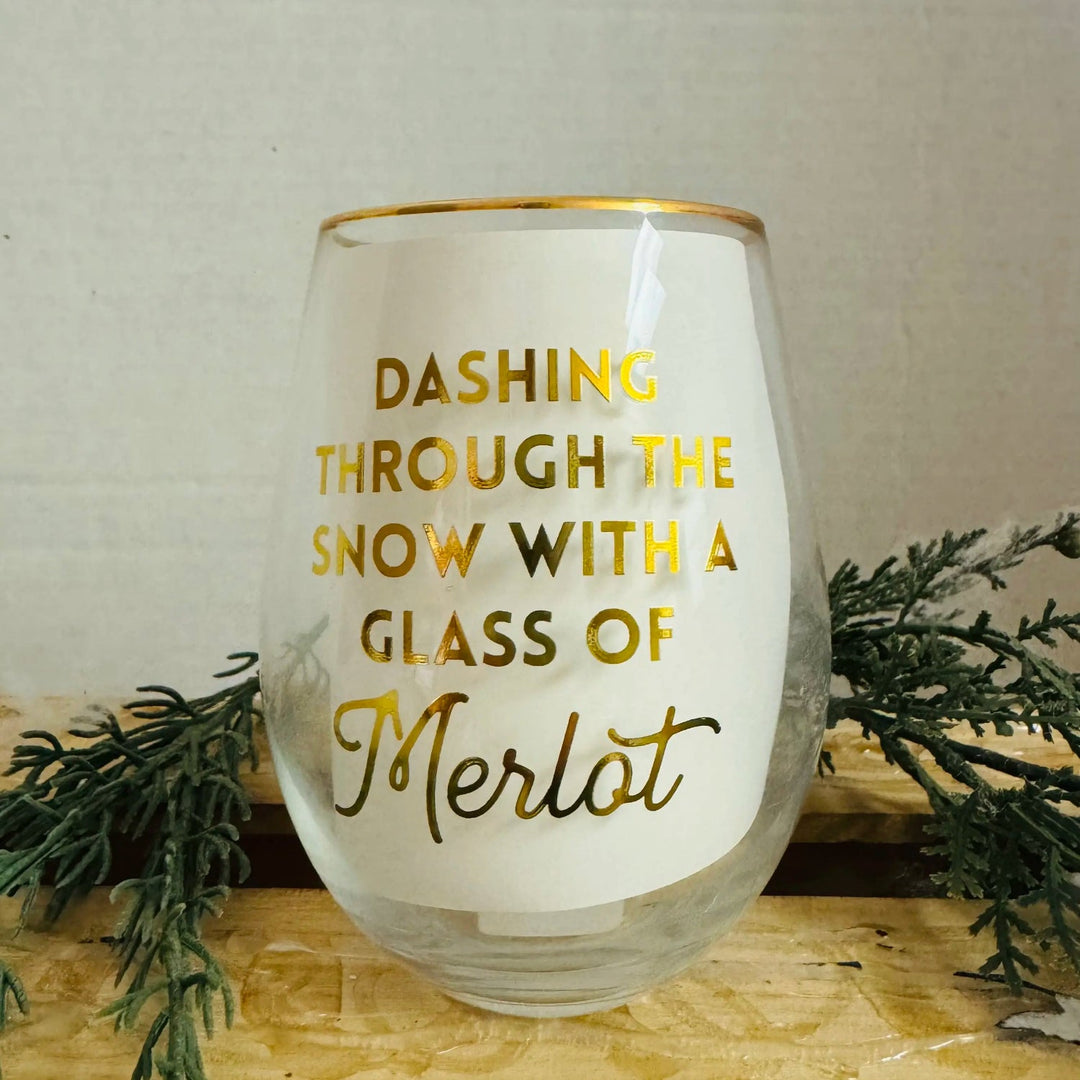 Stemless Merlot glass with a gold rim and festive gold text reading, “DASHING THROUGH THE SNOW WITH A GLASS OF Merlot.”.