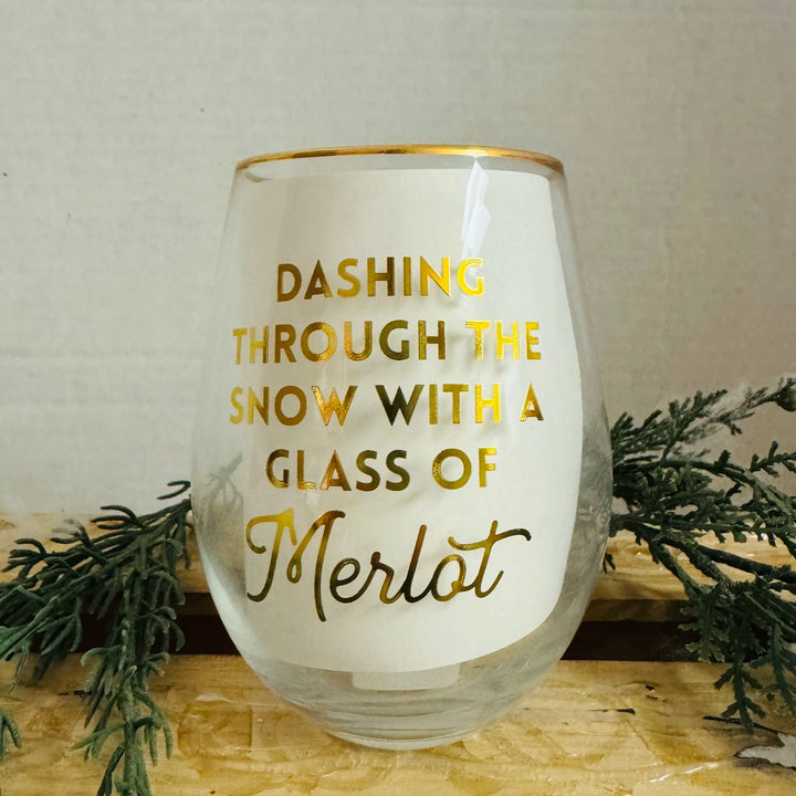 Stemless Merlot glass with a gold rim and festive gold text reading, “DASHING THROUGH THE SNOW WITH A GLASS OF Merlot.”.