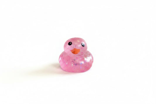Glitter Squishy Duck with a smiling face, sparkling pink finish, and orange beak. Available in small, medium, and large sizes.