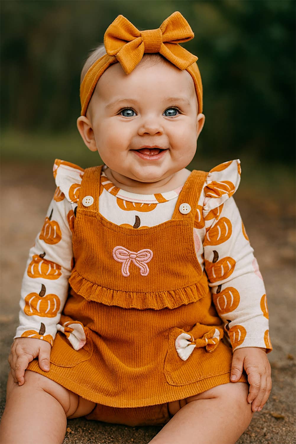 The Pumpkin Harvest 3-Piece Baby Dress Outfit includes a mustard yellow bow headband, pumpkin-print shirt, and terracotta corduroy jumper with bows, all made from soft, machine-washable cotton. Available in sizes 12-18M.