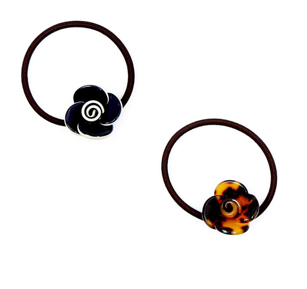 Two Camellia Hair Ties: one with a black rose-shaped accessory featuring a spiral center, and the other with a tortoiseshell-patterned rose showcasing a spiral center. Crafted from glossy material, attached to durable brown elastic bands. Available in one size.