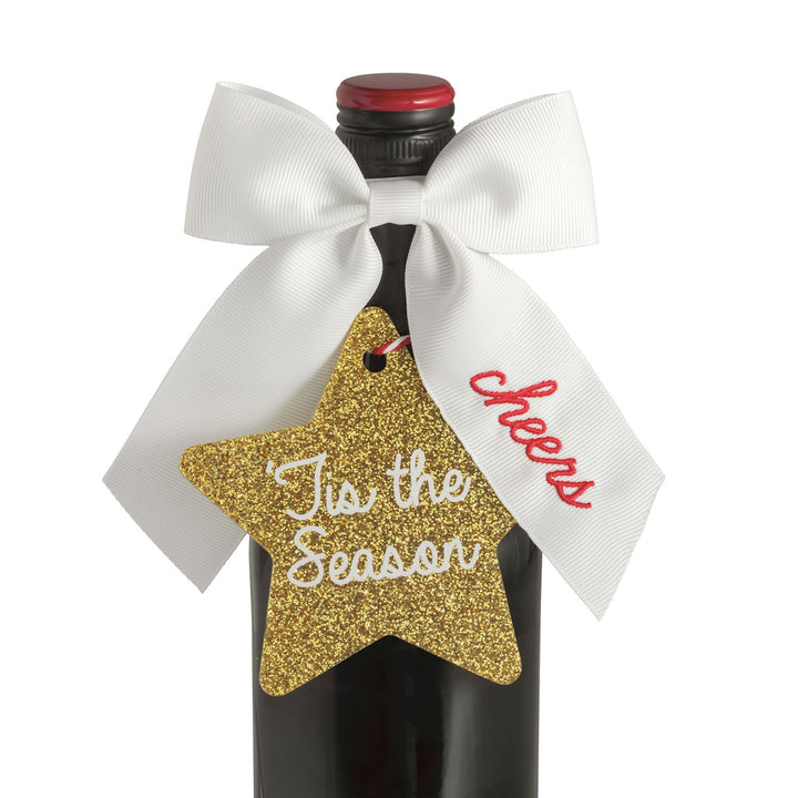 Bow Topper &amp; Gift Tag Set featuring a white ribbon and a gold glittery star-shaped tag printed with “’Tis the Season” and “cheers.”.