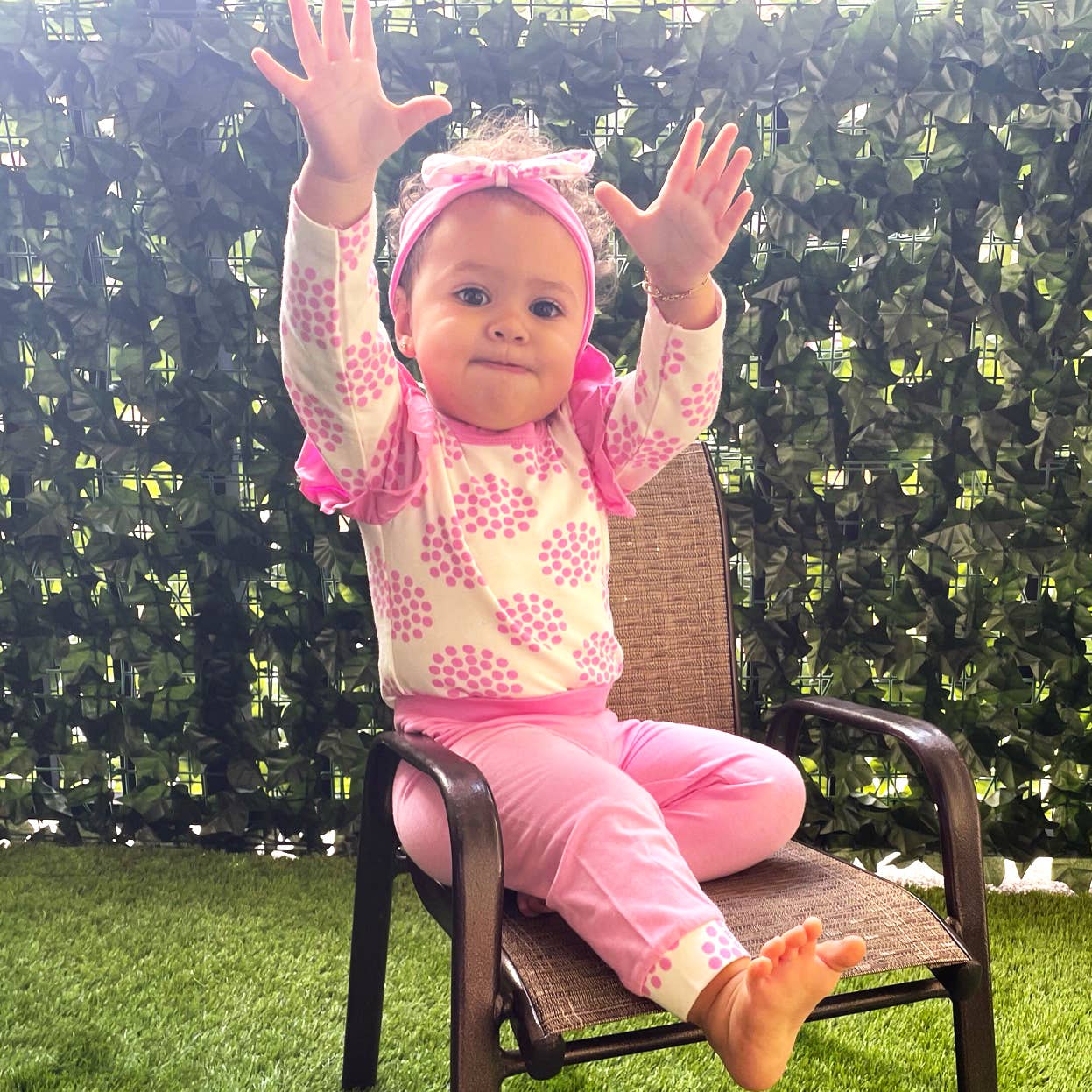 A toddler in a Layette Girls Baby clothes Shower Gift Pink Polka Dot Easter: 3-6 Mo with a matching headband sits on a patio chair, joyfully raising her hands up, against a backdrop of a lush green hedge.