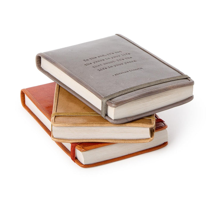 Handcrafted leather flip notebooks in brown, tan, and gray with elastic bands and embossed sayings. Available in multiple sizes.