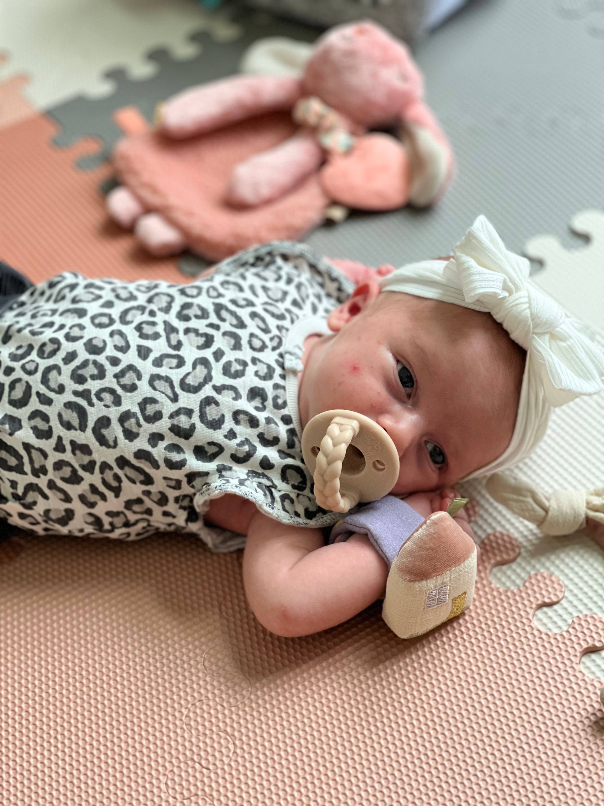 A baby wearing a leopard print outfit and a white bow headband lies on a play mat, chewing on an Itzy Bitzy Wrist Rattle: Cottage, with stuffed animals around.