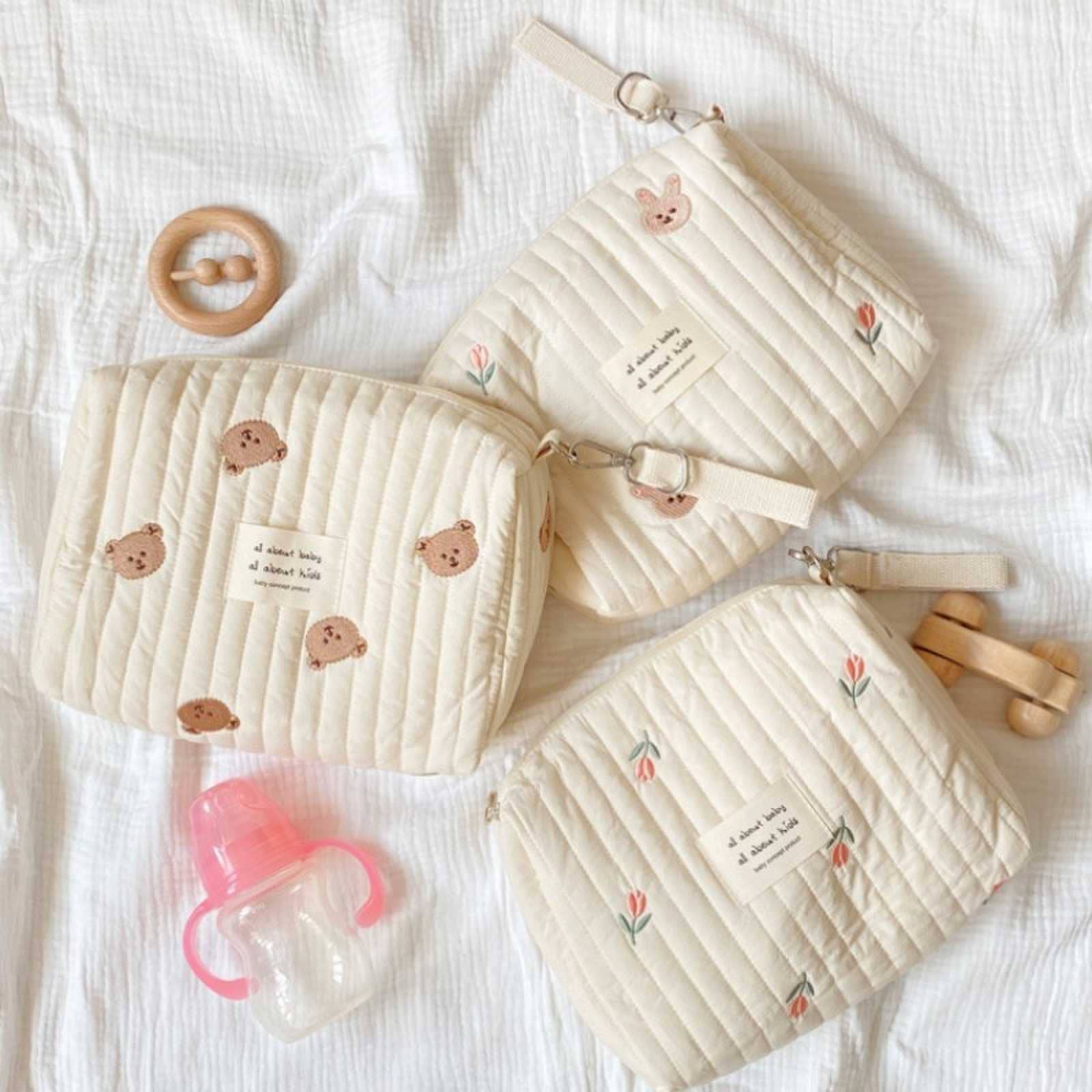 Three Little Bear Embroidered diaper bags with animal faces and small labels, made from high-quality durable materials, a wooden teething ring, and a pink pacifier, all arranged on a soft white fabric.