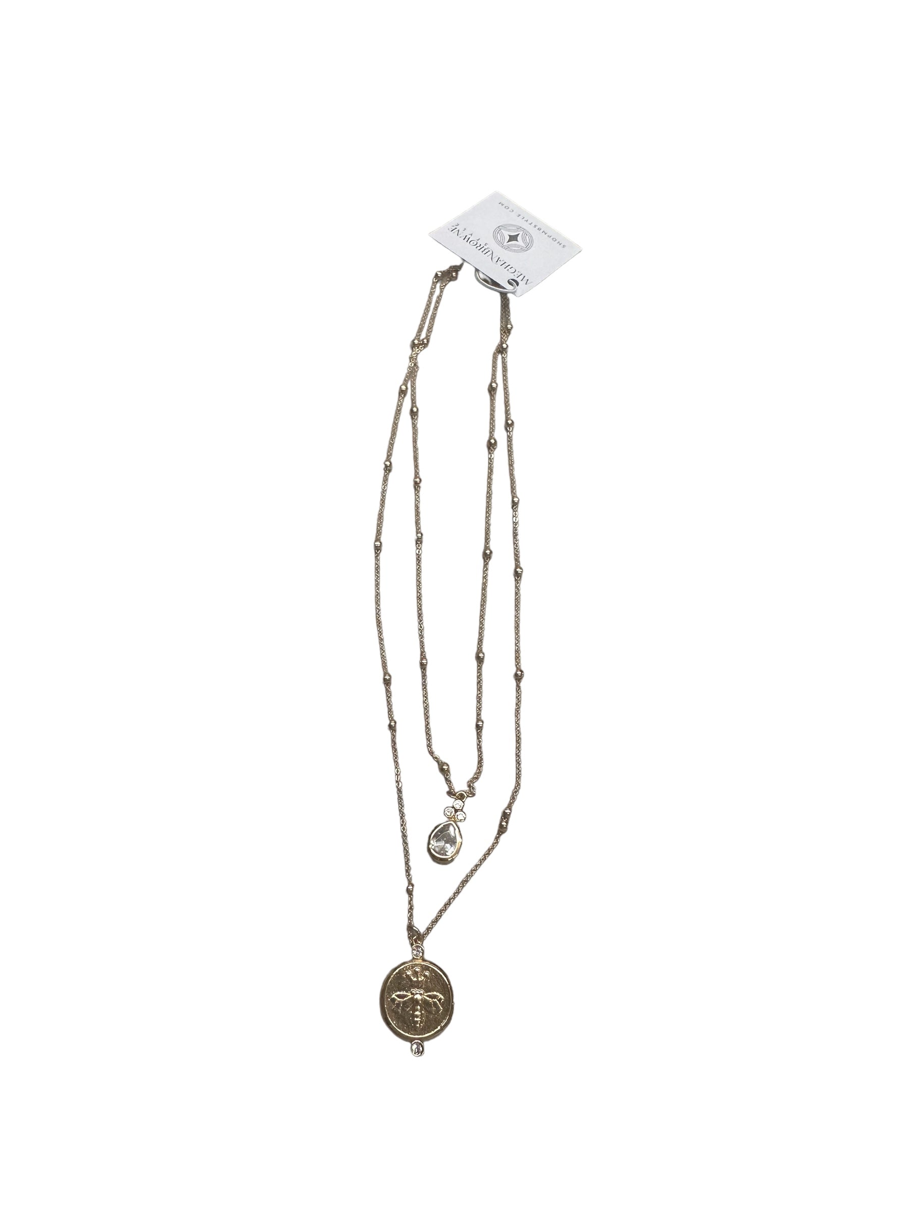 The Celery Gold 2 Chain Necklace features a double-strand design with delicate beads. The longer strand has a round medallion pendant with an intricate design, and the shorter strand has a small teardrop-shaped pendant. Available in one size.