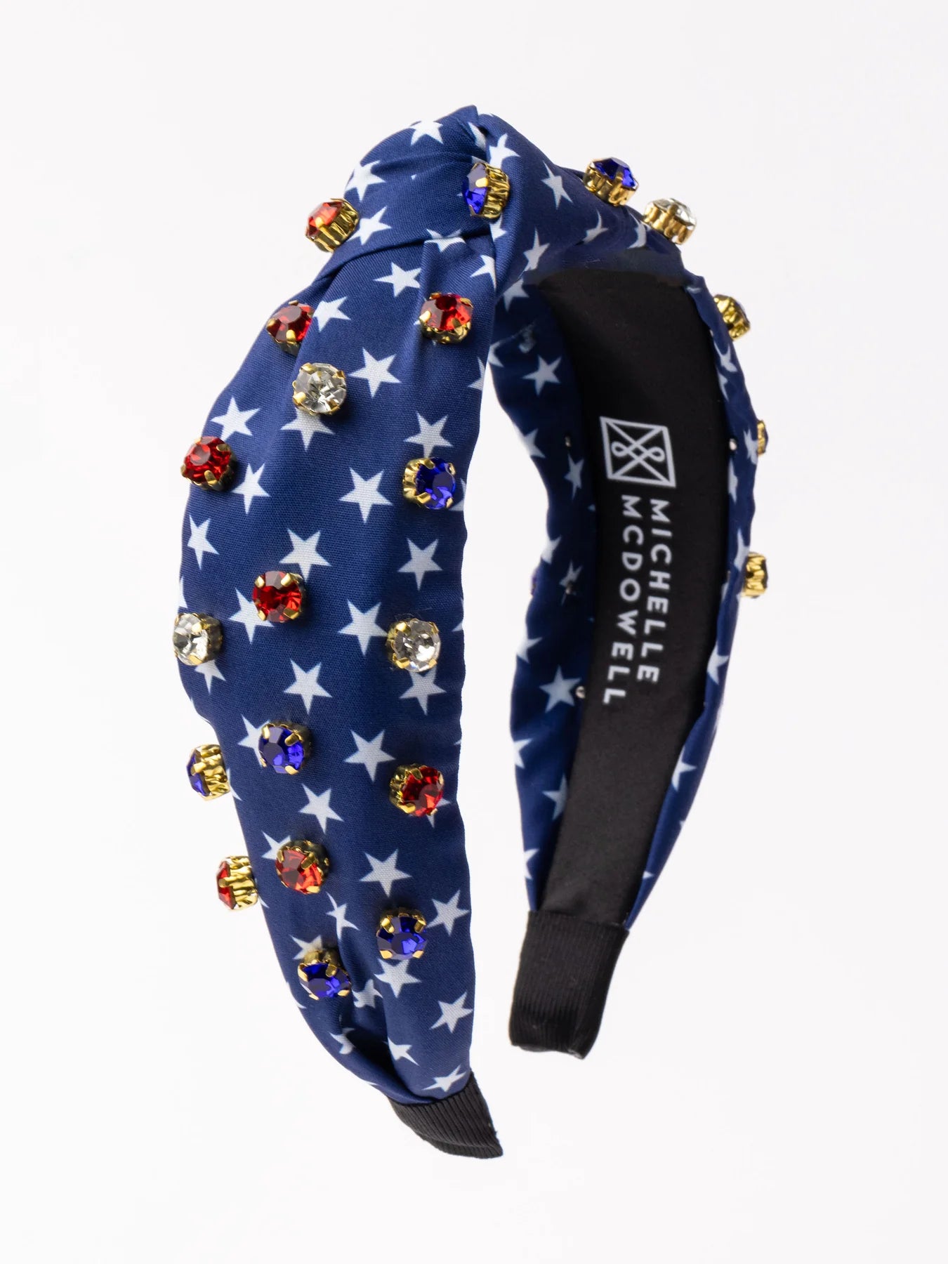 The product is an Americana Headband adorned with white stars and vibrant gem embellishments. It features a black segment displaying the text "michelle mcdowell". Available in one standard size.