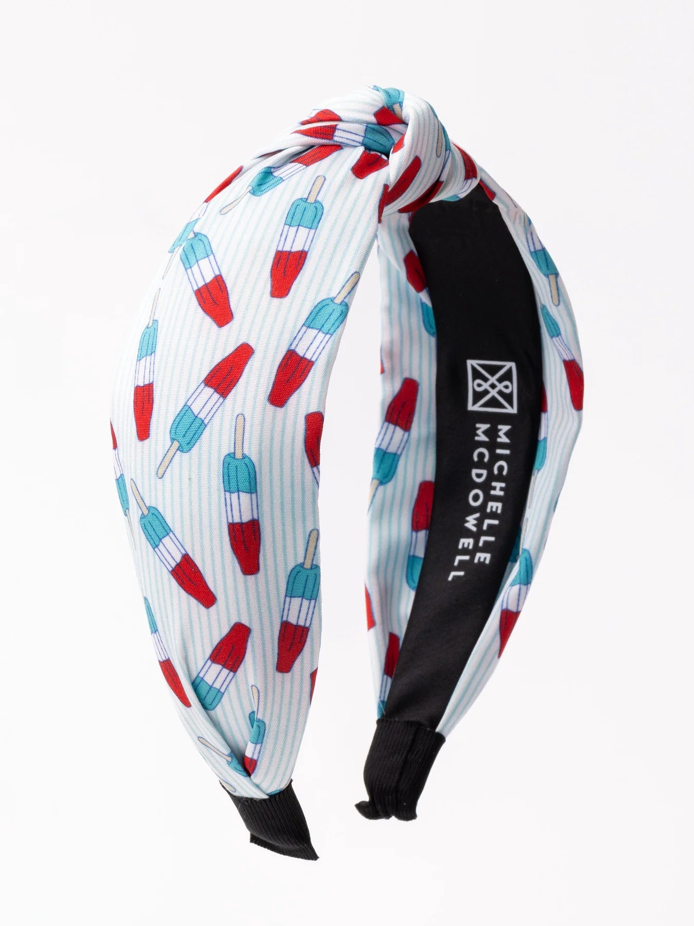 The product is a colorful Americana headband. It has a white background and features a repeating pattern of blue and red popsicles. The band includes a black interior label that bears the text "michelle mcdowell." Available in one-size-fits-all.