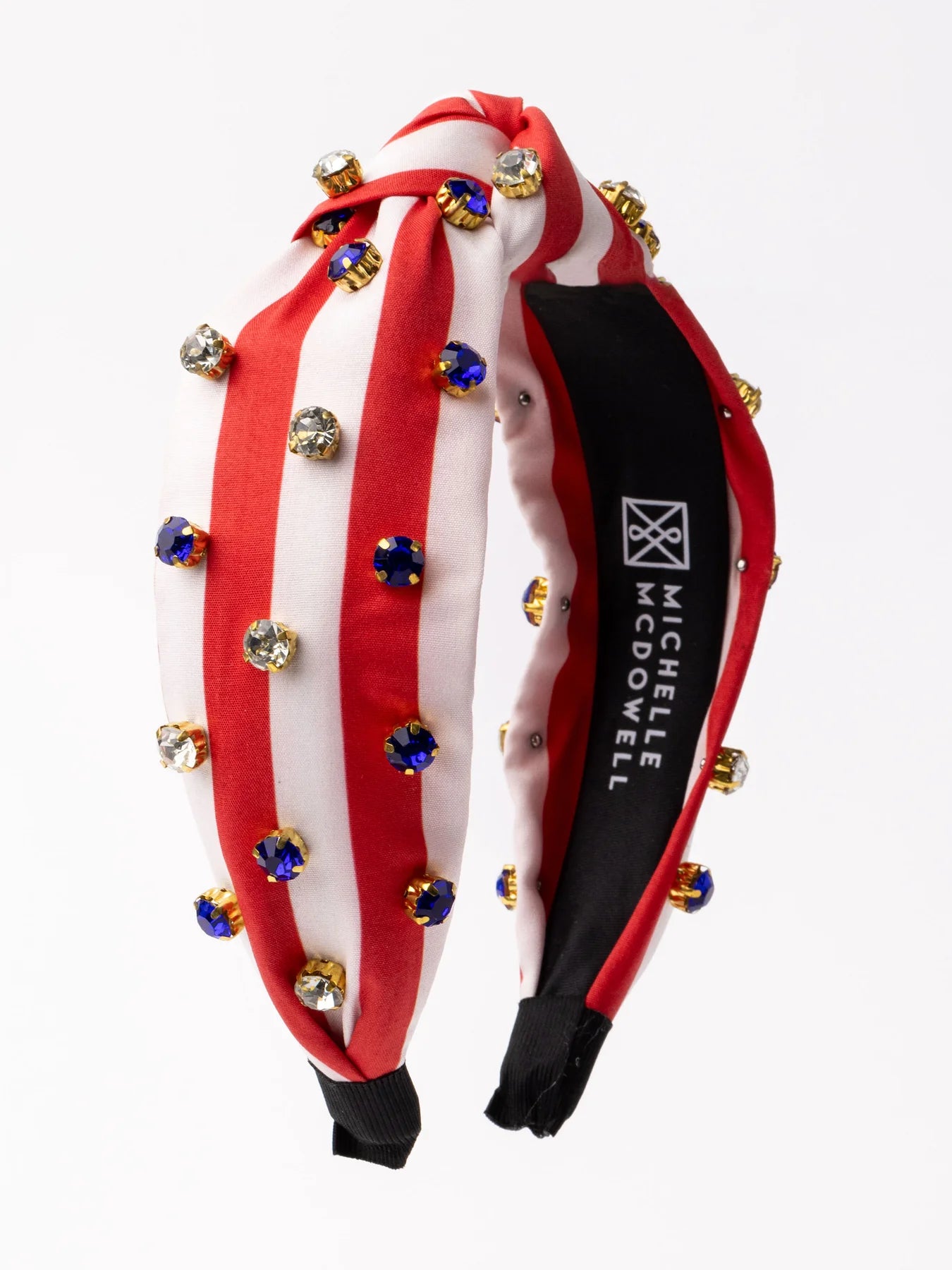 A red and white striped Americana Headband from the brand Michelle McDowell. It's decorated with golden baubles and blue gems, has a black interior lining stamped with the brand name. Available in one standard size.