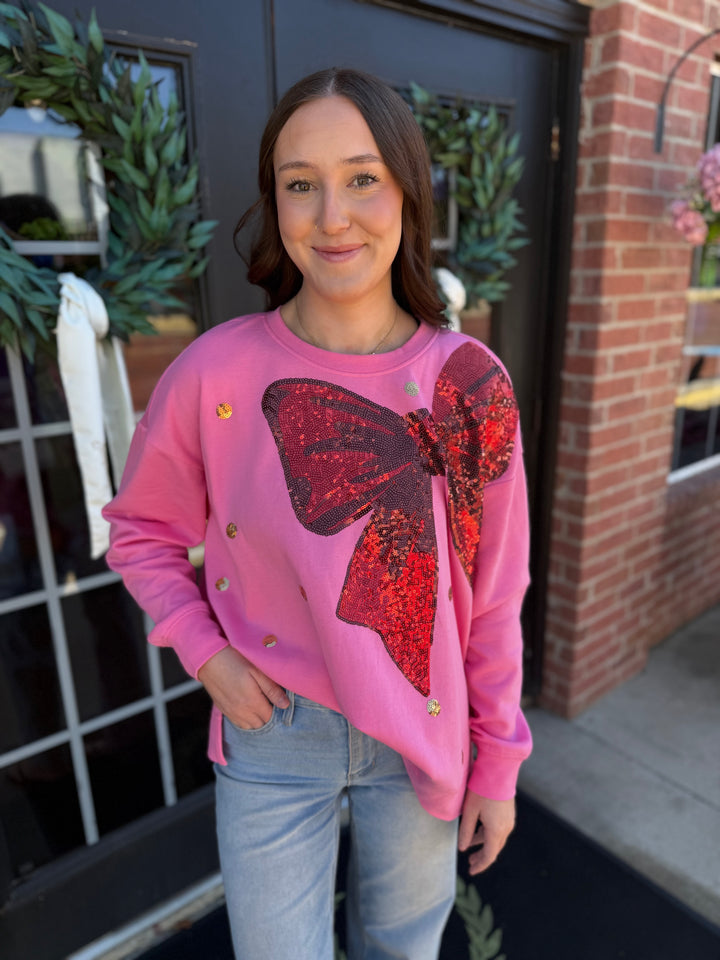 Pink sequin bow sweatshirt in a relaxed, long-sleeve cotton blend with a large, red sequined bow graphic. Sizes available: [Insert Sizes].