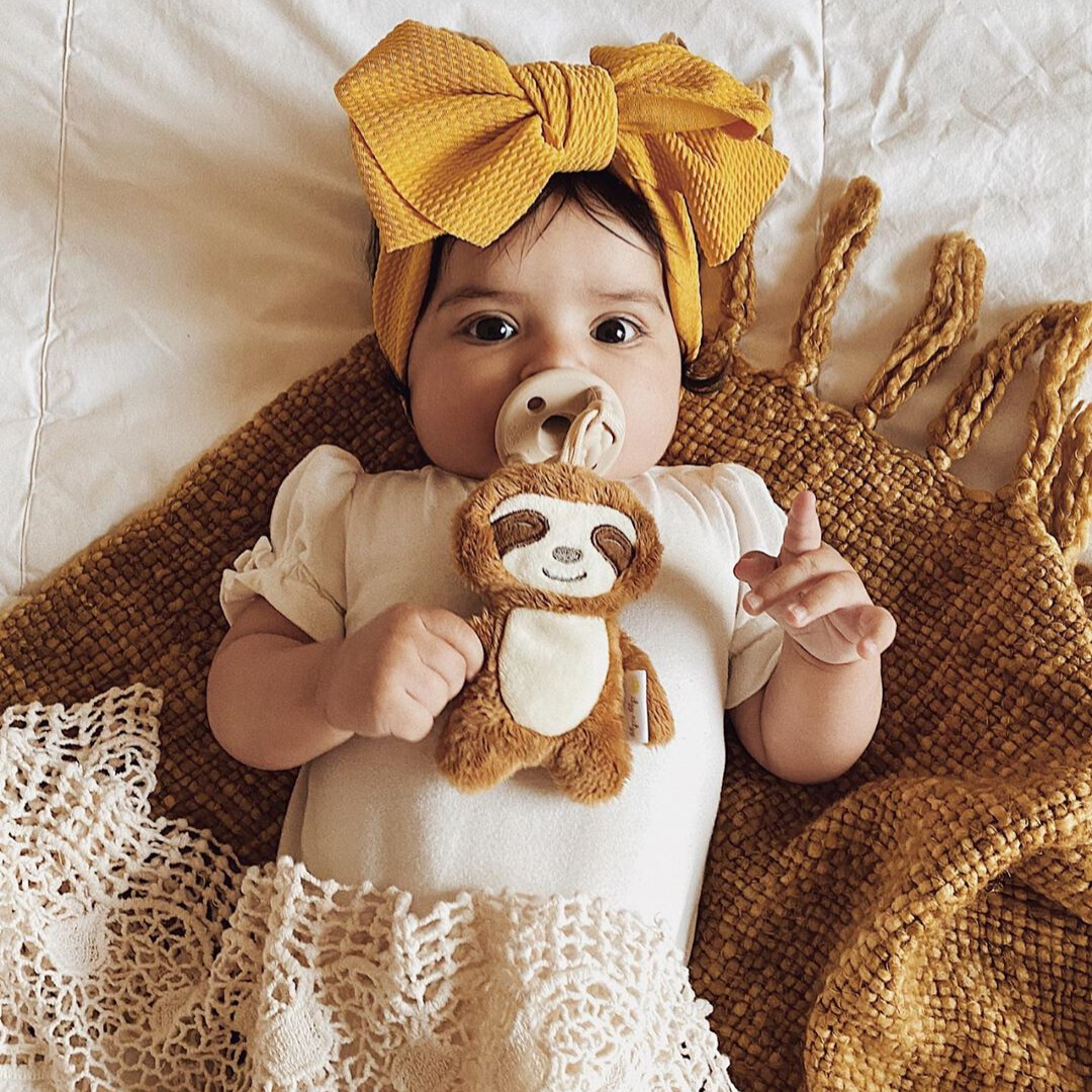 A baby with big eyes and a yellow bow headband lies on a cream blanket, holding a plush sloth toy and using a Sweetie Pal™ Plush & Pacifier: Dino.