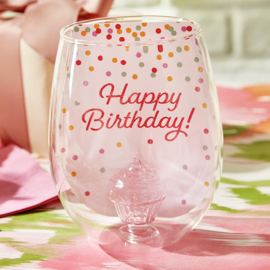 Celebration Cake Stemless Wine Glass" with "Happy Birthday!" in red and multicolored polka dots. Features a 20 oz. capacity and a glass cake inside the base. Durable glass on a pink and green surface. Available size: 20 oz.