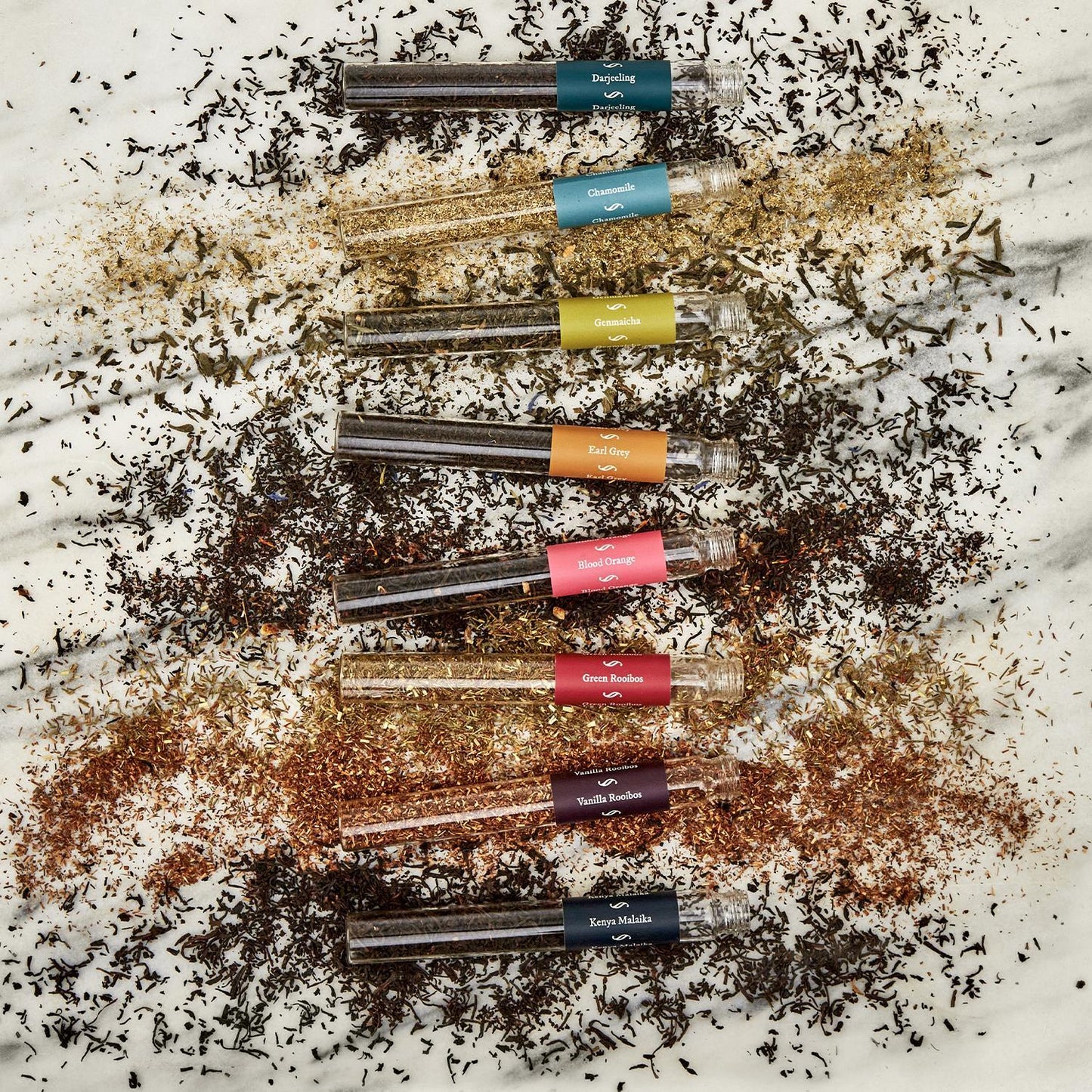 Eight vibrant test tubes filled with assorted teas, presented diagonally on a marble surface. Loose leaves are scattered around open tubes labeled with different flavors. Comes in a gift box.