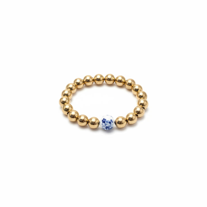 The Chinoiserie Pearl Stretch Bracelet has shiny gold beads with a single white bead featuring blue floral patterns. Available sizes: Small, Medium, Large.
