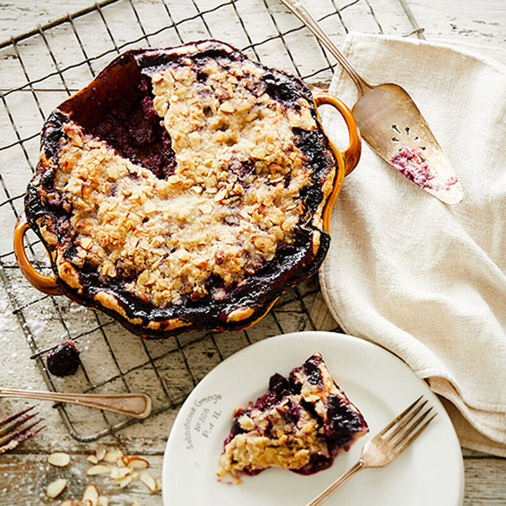 Baked berry crumble pie with a missing slice on wire rack; available in whole pie and individual slices.