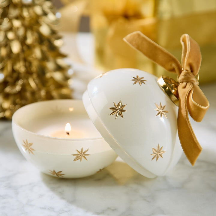 Sparkling Rose Ornament Candle in an open white ceramic design with gold star motifs, tan velvet ribbon, and a lit wick.