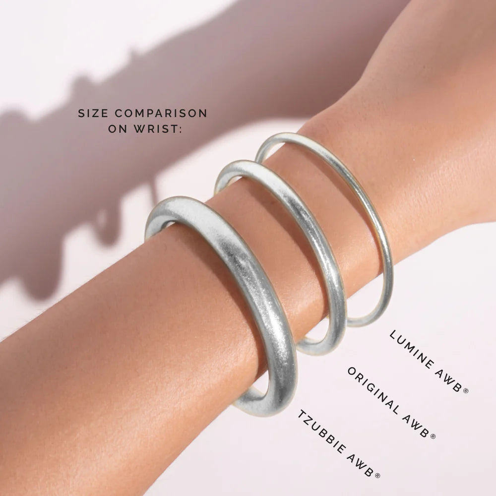 Three silver AWB bangles—Lumine (Large), Original, and Tzubbie—shown side by side for size comparison; text reads: “Size comparison on wrist:”. Sizes available: Large (Lumine), Original, Tzubbie.