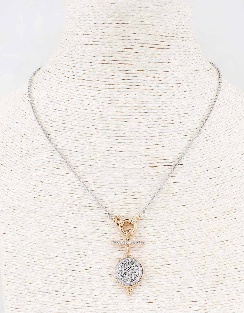 The Silver & Gold Cove Necklace features a silver chain, gold toggle clasp, and round textured silver pendant. Available in 16″, 18″, and 20″ lengths.